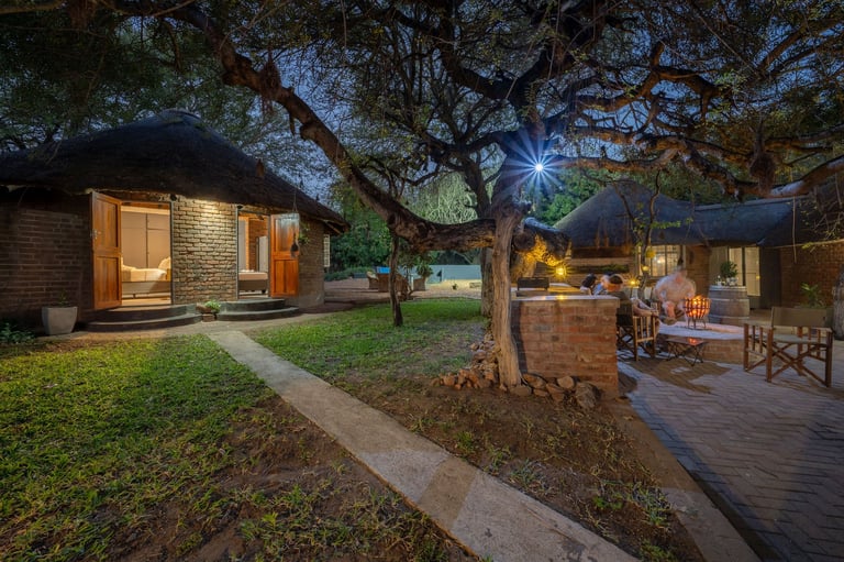 Twilight view of a modern residential courtyard with illuminated brick cottage, deck seating area, and mature trees on green lawn