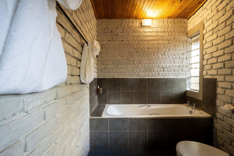 A brick bathroom with a bathtub surround in dark tile, white towels on wall, yellow ceiling light, and partial view of white toilet