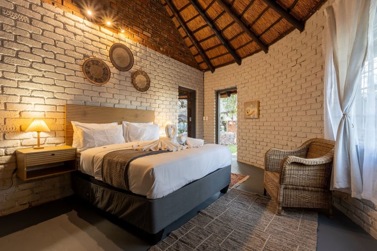 Modern bedroom with exposed brick walls, wooden ceiling beams, queen bed with white bedding, bedside lamp, woven wall baskets, and rattan chair by window