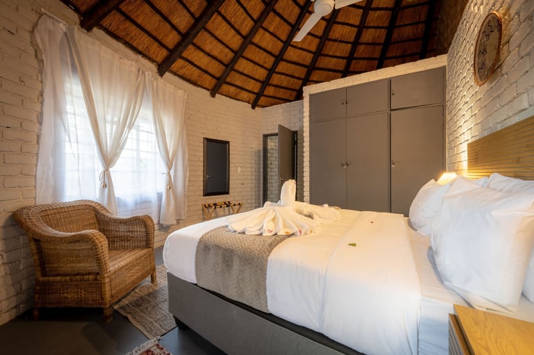 Spacious modern bedroom with white brick walls, wooden roof beams, twin beds with white bedding, wicker chair, and tall wardrobe