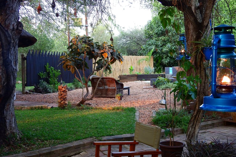 Backyard garden at dusk with mature trees, wooden benches, fire pit, and blue lantern among plantings