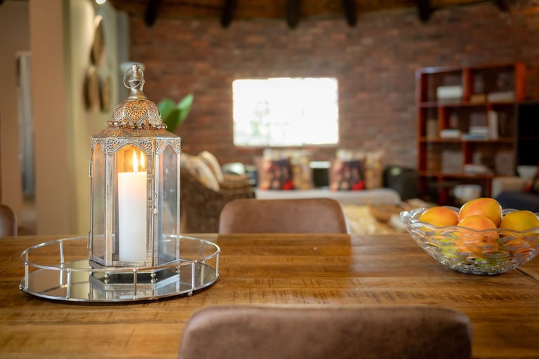 Wooden dining table with a lit candle lantern, fruit bowl, and living room with brick wall and TV in the background