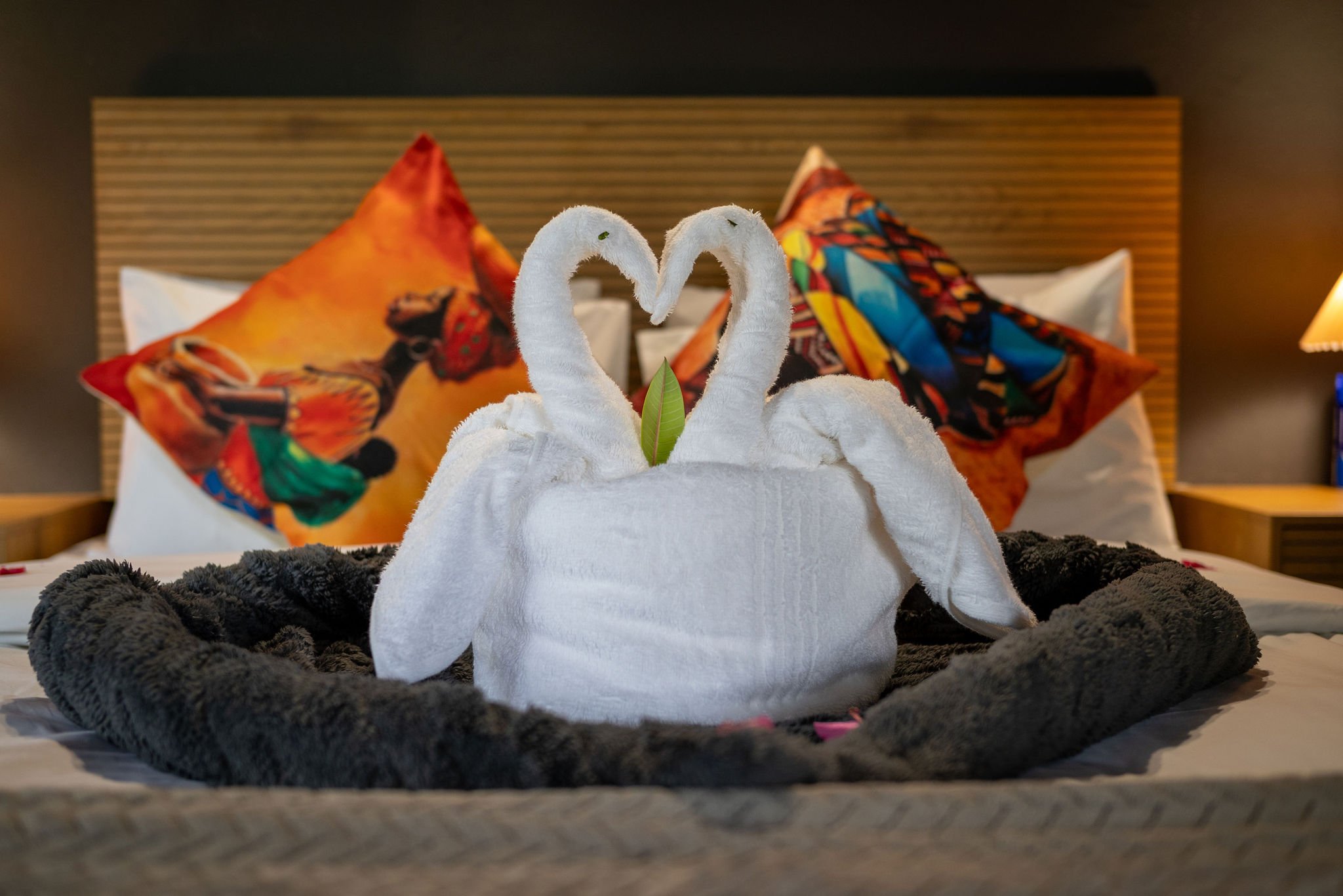 Two rolled towels shaped into swans with heads forming a heart, sitting on a bed with colorful pillows in the background