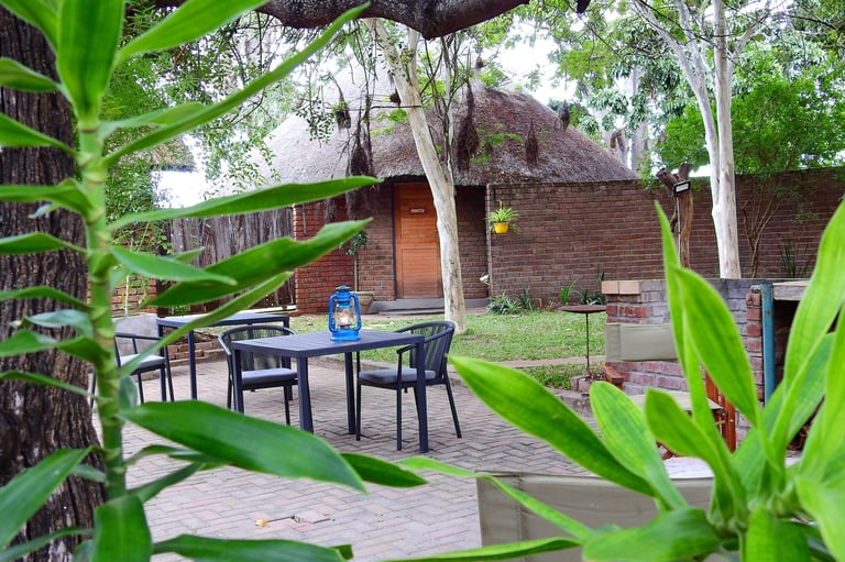 Outdoor patio with dining table and chairs beneath trees, rustic brick buildings visible in background surrounded by green vegetation