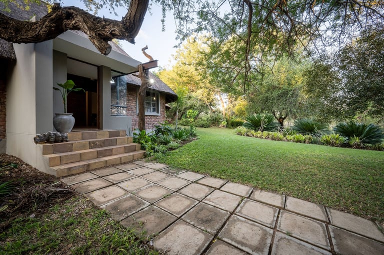 Modern home exterior with stone patio, entrance walkway, large lawn, and mature trees providing shade