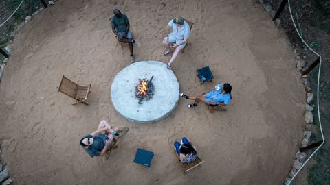 Aerial view of six people sitting around a circular fire pit on sandy ground at dusk