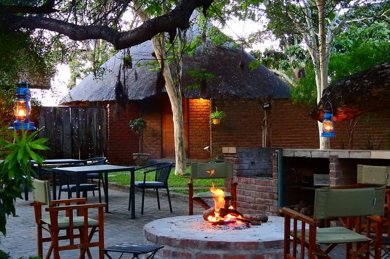 Evening outdoor patio with fire pit, wooden chairs, dining tables, and thatched-roof structures surrounded by trees
