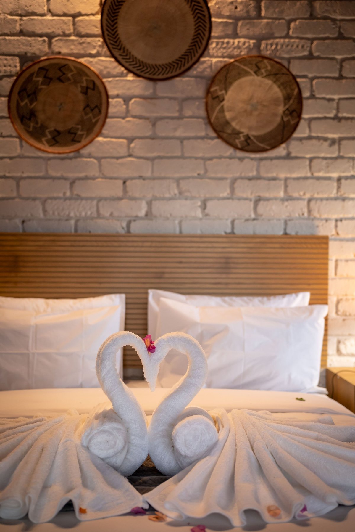 Two white towels arranged in heart shape on bed with white pillows and decorative plates on brick wall