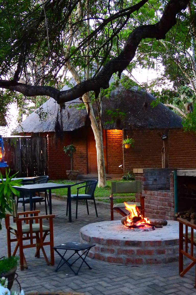 Backyard patio with fire pit, dining table, and wooden cabins under a large tree at dusk