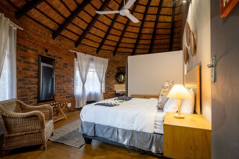 Modern bedroom with exposed brick walls, thatched ceiling, wicker chair, and queen bed with white linens